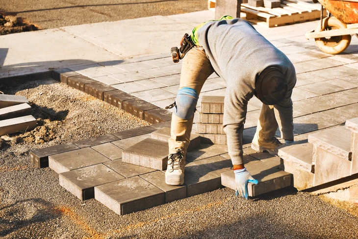 Ouvrier en tenue de travail posant des pavés sur lit de sable, utilisant un maillet à tête blanche pour ajuster l'alignement