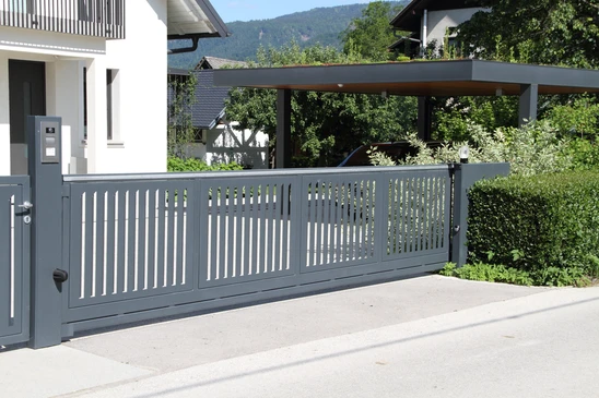 Sc&egrave;ne ext&eacute;rieure r&eacute;sidentielle avec cl&ocirc;ture en bois blanc &agrave; lames verticales espac&eacute;es, piliers ma&ccedil;onn&eacute;s beiges, v&eacute;g&eacute;tation luxuriante en arri&egrave;re-plan incluant arbres et arbustes verts